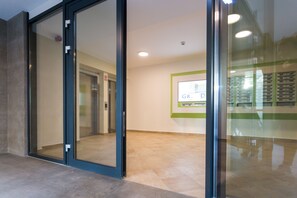 Interior moderno com portas de vidro, elevador visível e painel informativo fixado na parede.