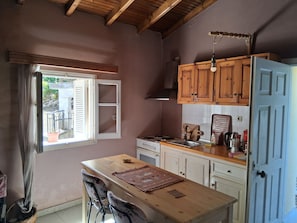 A kitchen with wooden cabinets, a stove, and a window with curtains. - Elm Tree tiny but unique property in Pantokratoras mountain :) (Corfu)