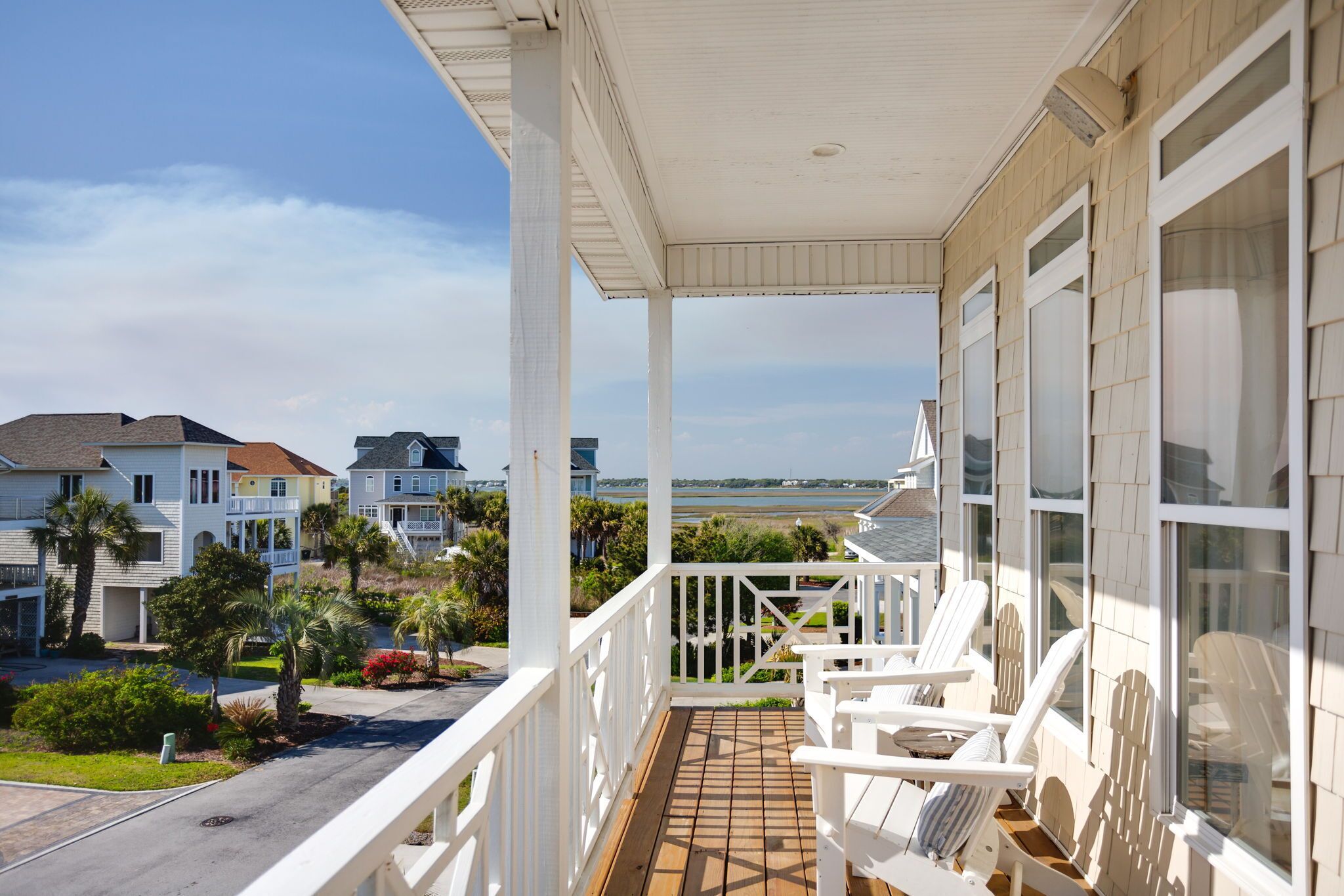 Een houten terras met witte leuningen en stoelen, met uitzicht op een woongebied met huizen en een strand.