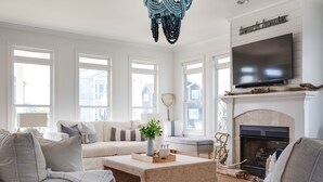 A living room with a fireplace, a television mounted above it, and a large window with a view of houses. - The Big Dream Getaway 💙Private Heated Pool Sleeps 13 (Atlantic Beach)