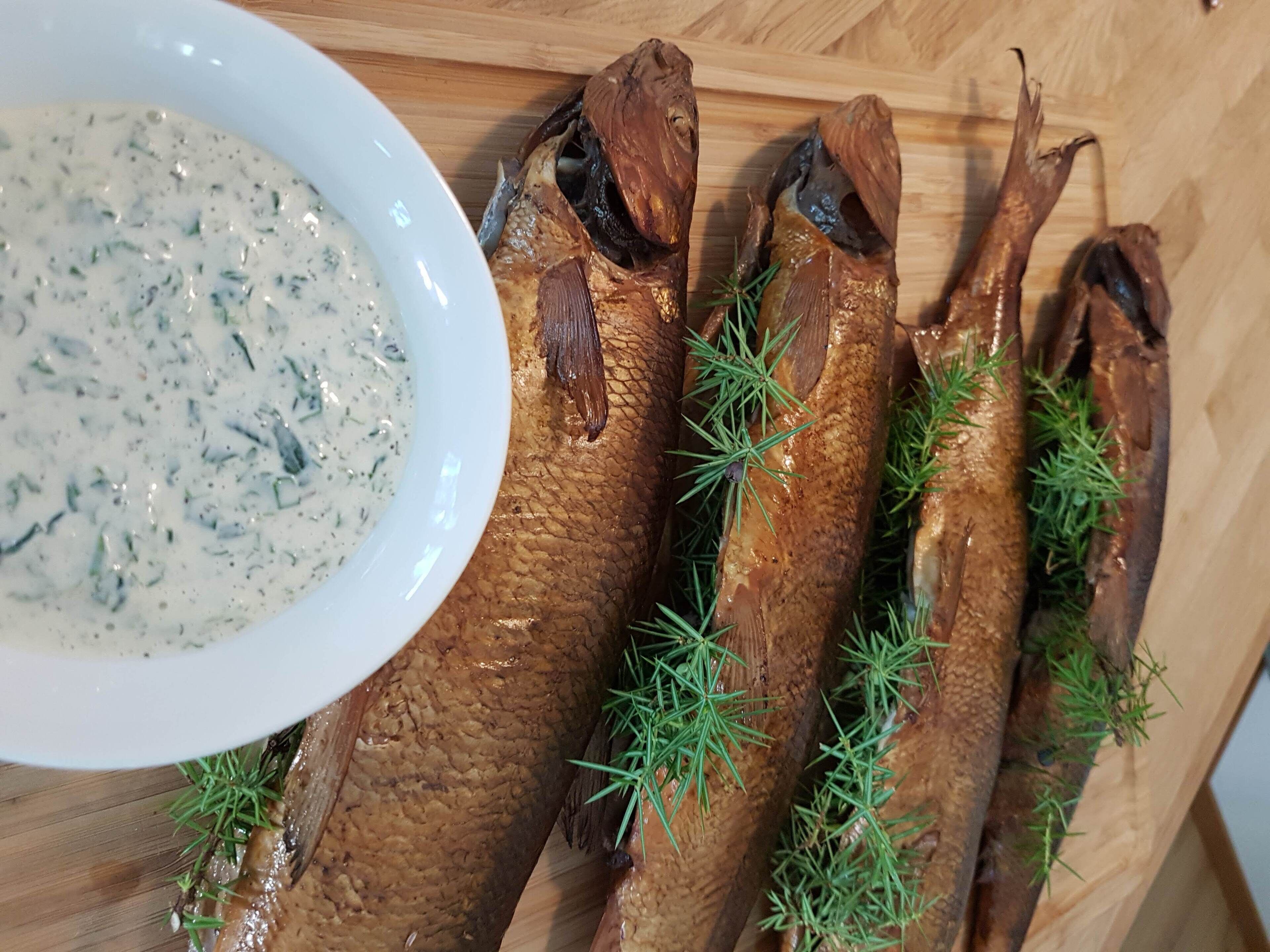 A wooden cutting board with several smoked fish and a bowl of sauce.