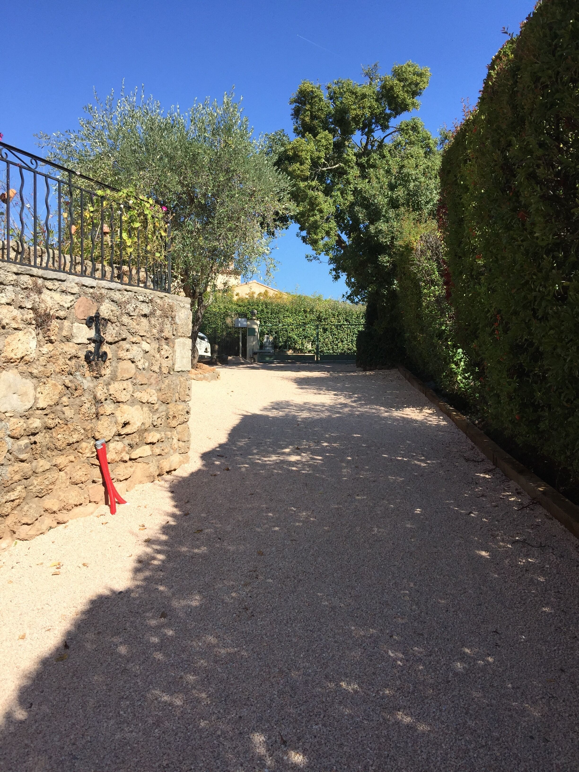A gravel driveway leading to a stone wall with a metal gate, surrounded by greenery and trees.