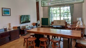 A spacious room with a long wooden table, several wooden stools, a sofa, a television, and a large window with curtains. - Clove House (Hengchun)