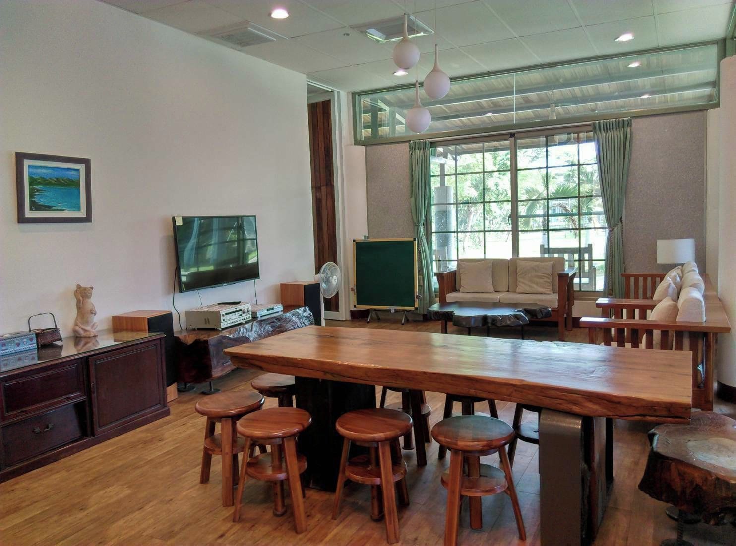 A spacious room with a long wooden table, multiple stools, a television, a green chalkboard, and a large window with curtains.