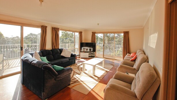 A spacious living room with a dark sofa, beige armchairs, a flat-screen TV, and large windows offering a view of the outdoors.