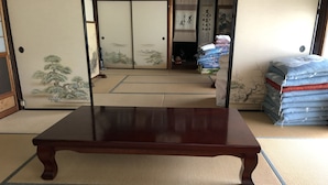 A traditional Japanese room with tatami flooring, a wooden table, sliding doors with artwork, and a stack of bedding in the background. - Hidejiro (Fukuchiyama)