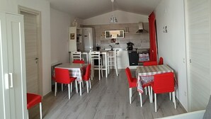 Eine kompakte Küche mit weißen Schränken, einem Kühlschrank und einer Mikrowelle. Zwei Esstische mit roten Stühlen und eine Couch im Vordergrund.