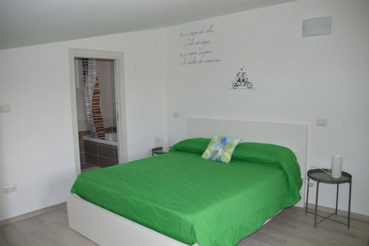 Une chambre avec une couvre-lit verte, une tête de lit blanche, un motif décoratif représentant un vélo sur le mur et une petite table de chevet avec une lampe.