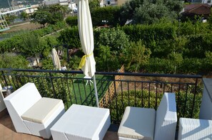 Ein Balkon mit weißen Korbmöbeln, einem gelben Sonnenschirm und Blick auf einen Garten sowie Gebäude.