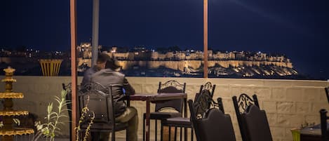 A person seated at a table with a view of a fortified hilltop structure in the background.