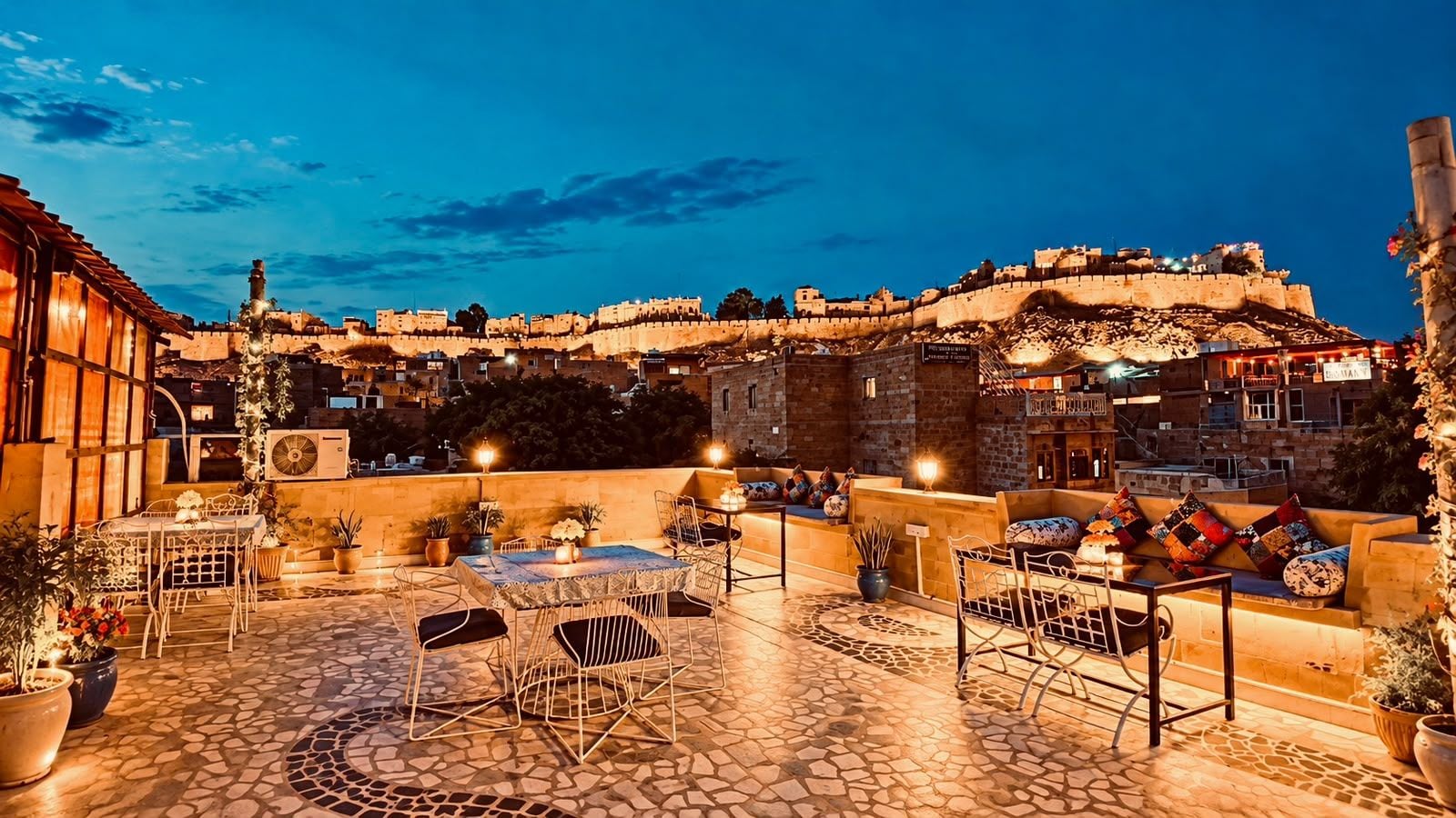 Un patio exterior con mesas y sillas, vista a una muralla histórica y cielo despejado.