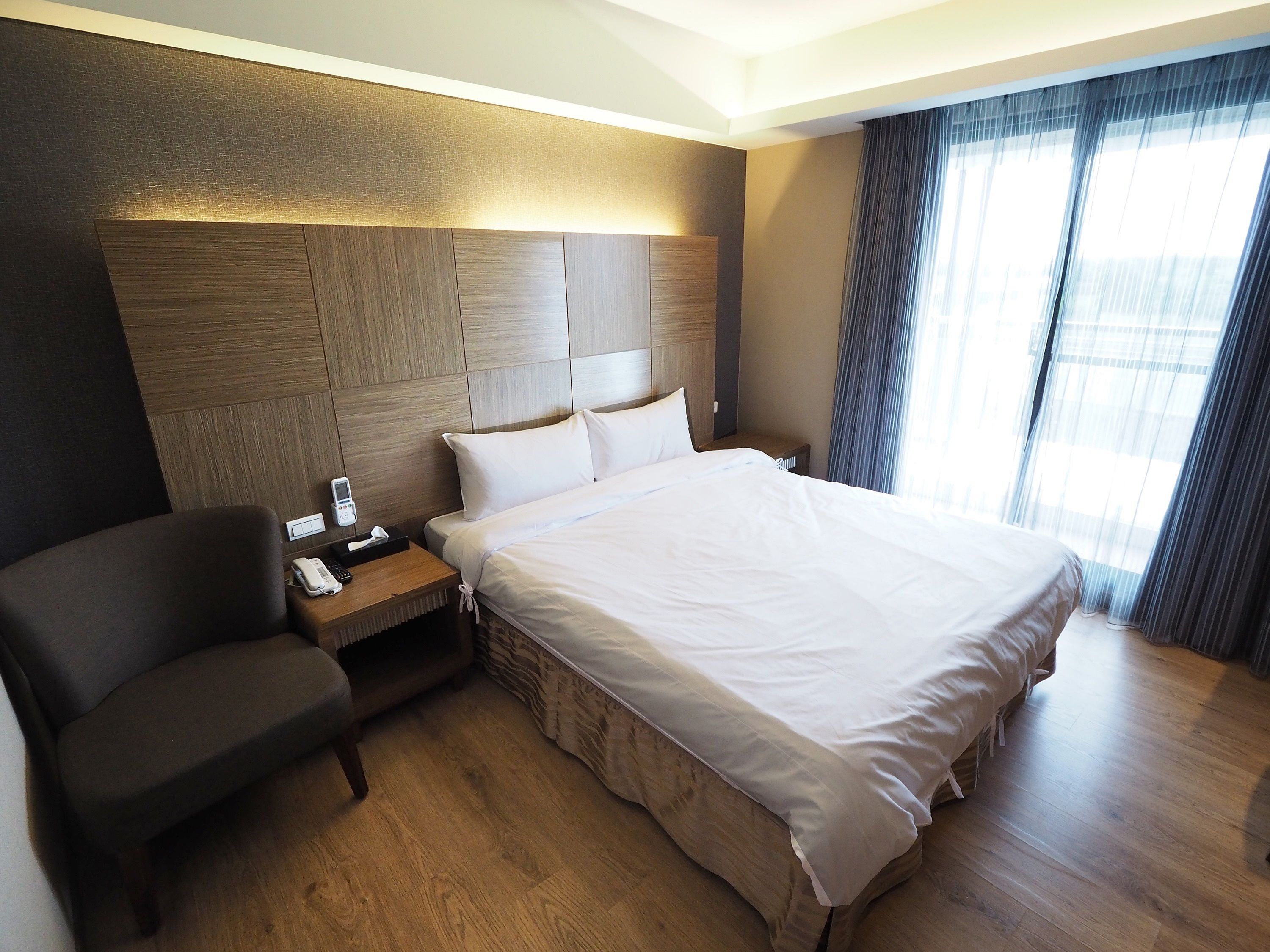 Habitació d'hotel amb un llit gran, una taula de nit amb un telèfon, una cadira i un panell d'il·luminació al capçal del llit.