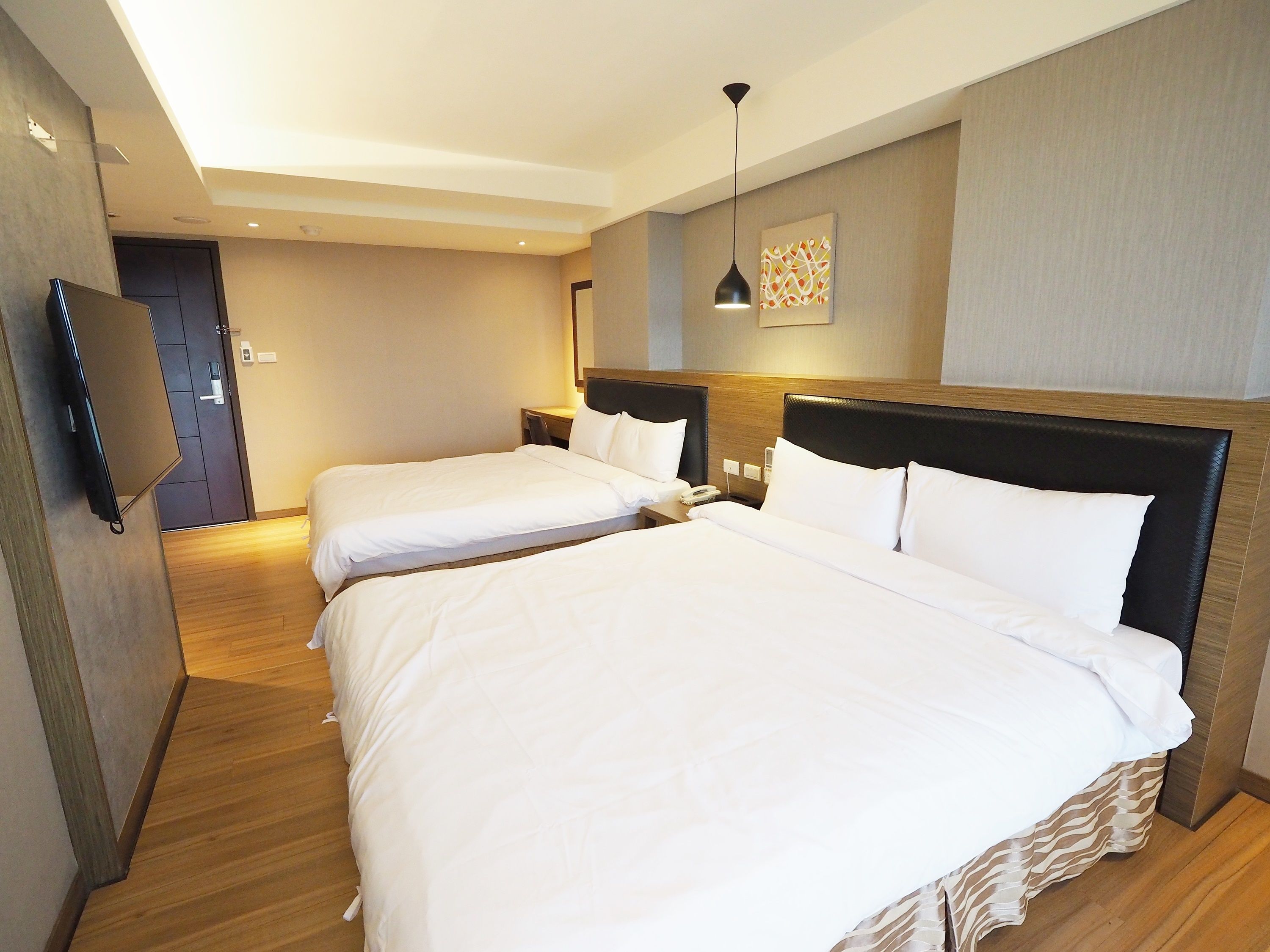 A hotel room with two beds, a TV mounted on the wall, and a modern light fixture.