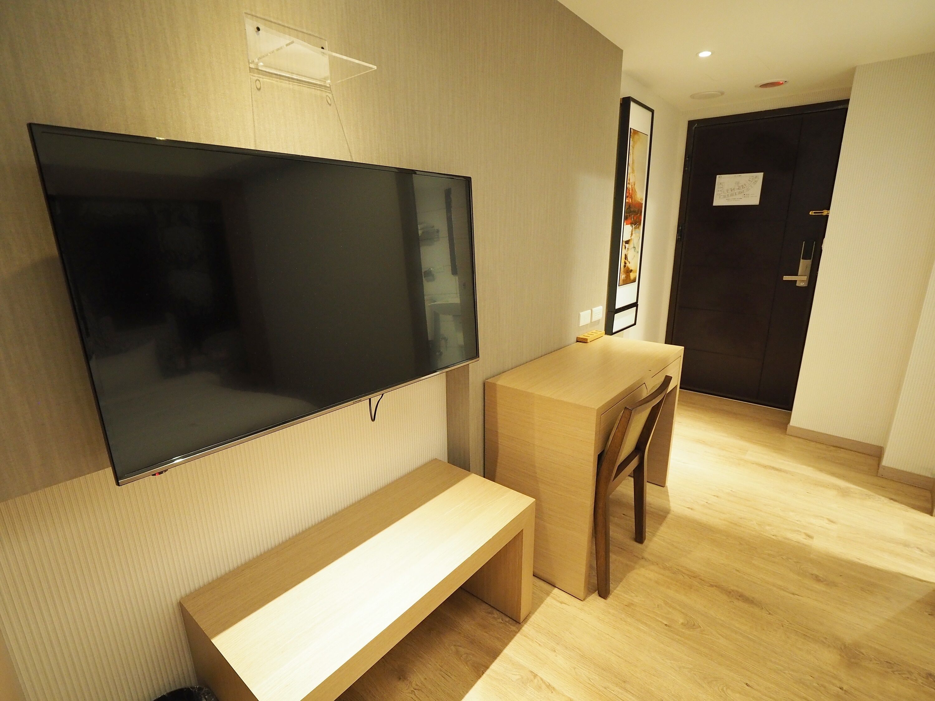 A modern hotel room with a large flat-screen TV mounted on the wall, a wooden desk, a bench, and a door with a framed picture.