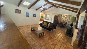 Een ruime woonkamer met een hoge plafond, zichtbare houten balken, een zwarte bank met gele kussens, een houten salontafel en een open haard.