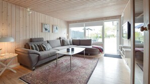 Un salon moderne avec un canapé d’angle gris, une table basse ronde et un tapis à motifs. Deux œuvres d’art encadrées ornent le mur, ainsi qu’une lampe sur pied et un téléviseur fixé au mur.