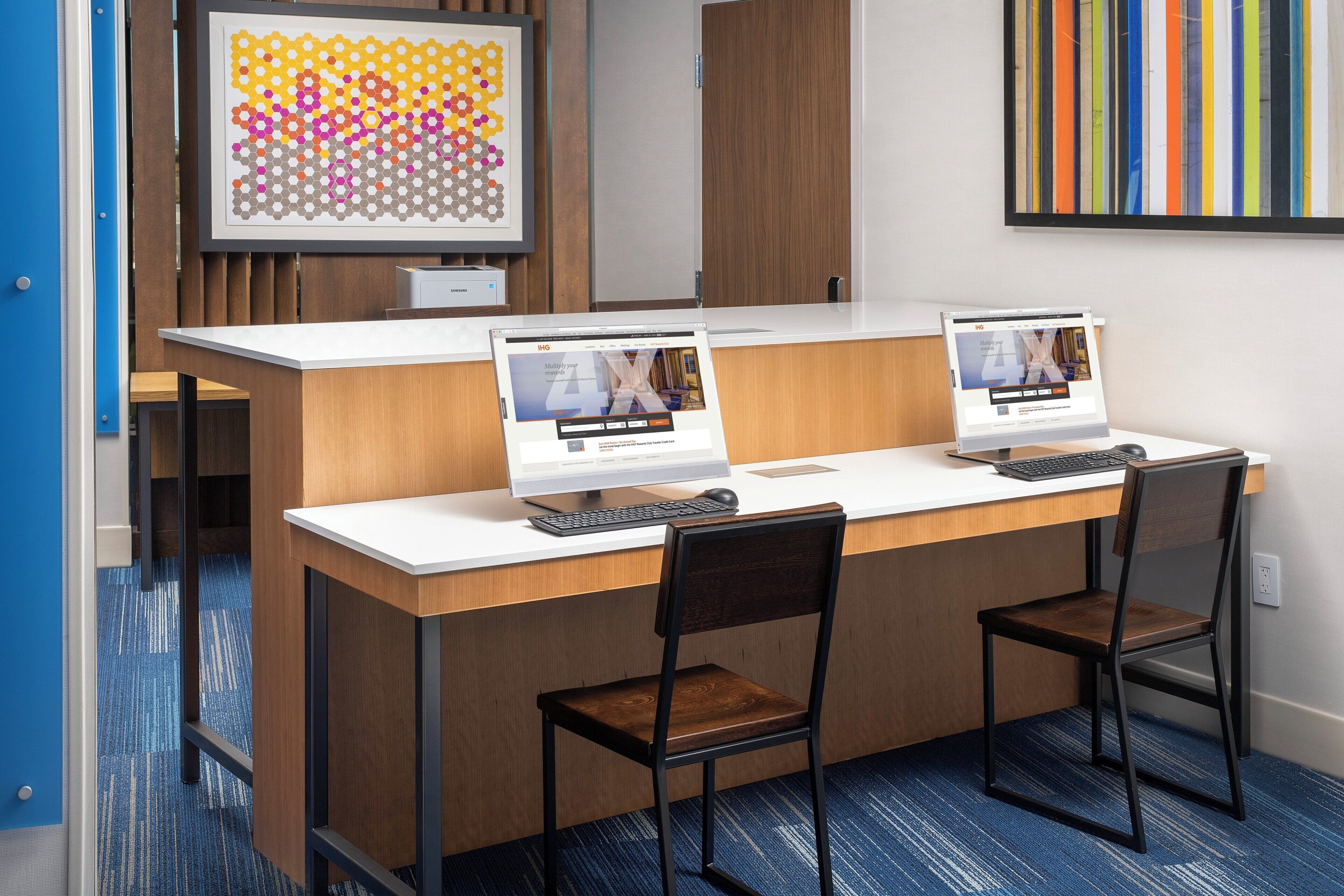 Un mostrador de recepción con dos estaciones de trabajo de computadora, una impresora Samsung y una gran obra de arte abstracto en la pared.