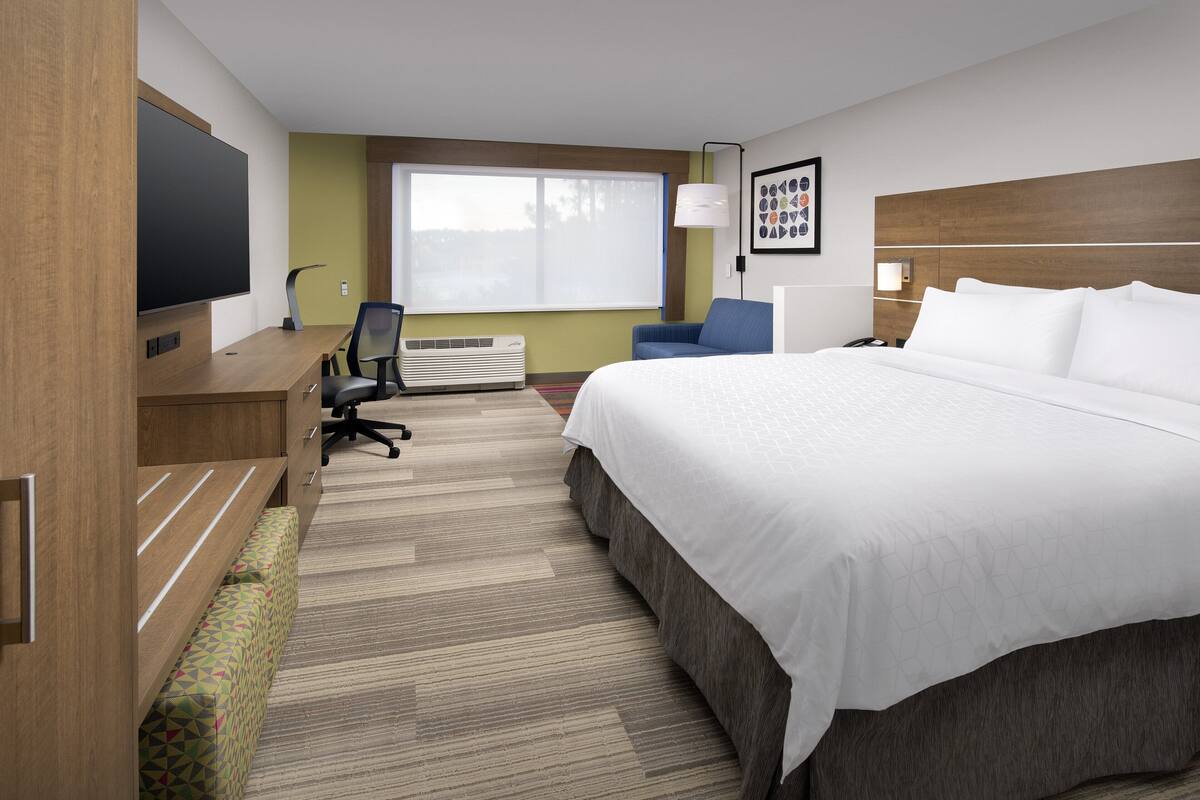 A hotel room with a large bed, a desk with a flat-screen TV, a chair, a bench, and a window with blinds.