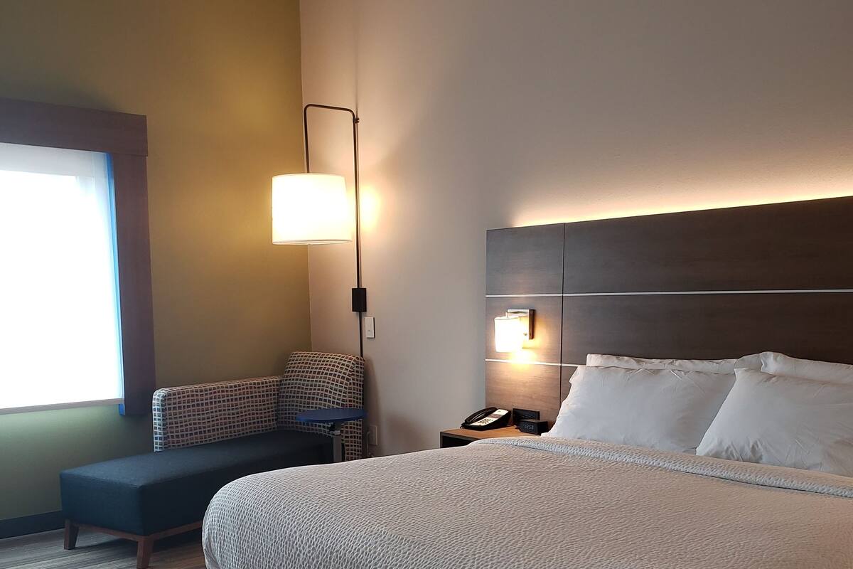 A hotel room with a large bed, a bedside table with a lamp, a chair, and a window with blinds.