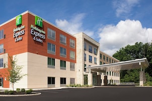 Um hotel Holiday Inn Express & Suites com design moderno, apresentando um logotipo verde e branco marcante, uma entrada coberta e um estacionamento.