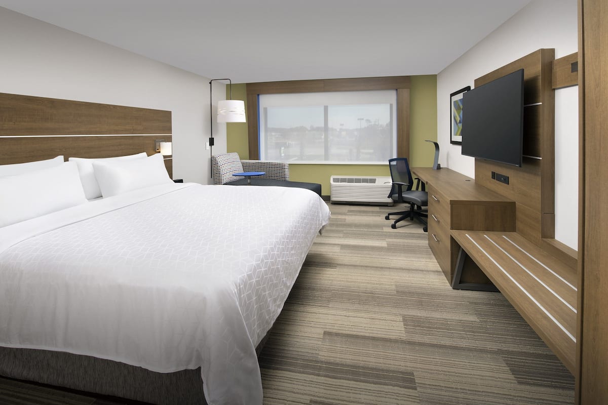 A hotel room with a large bed, a desk with a chair, a flat-screen TV, and a window with a city view.