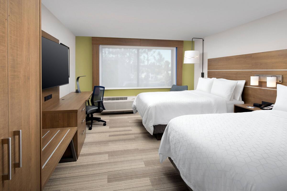 A hotel room with two beds, a desk with a chair, a flat-screen TV, a window with blinds, and a wall-mounted lamp.