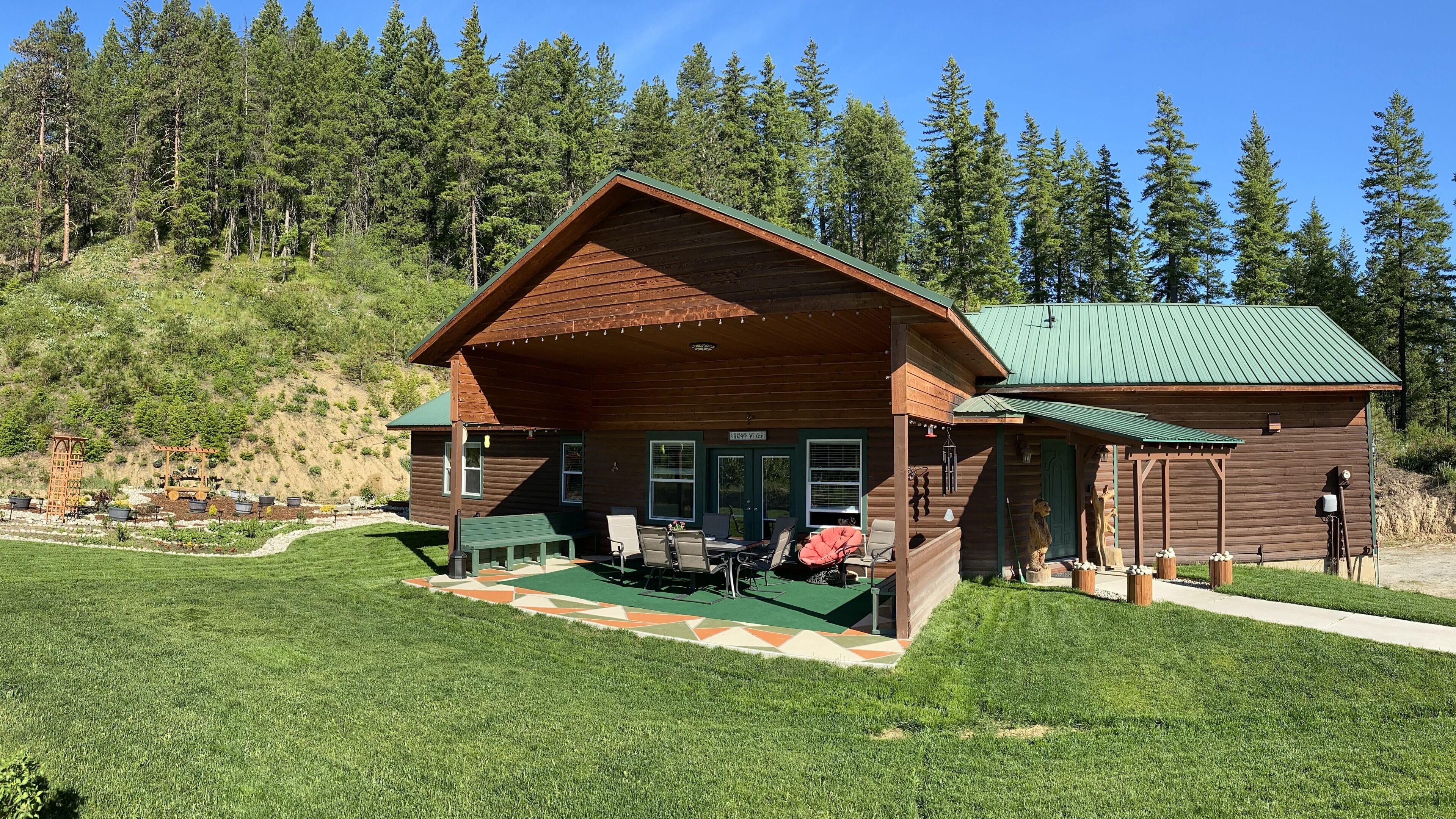 Una baita di legno con tetto verde, un portico con sedie e un piccolo giardino.