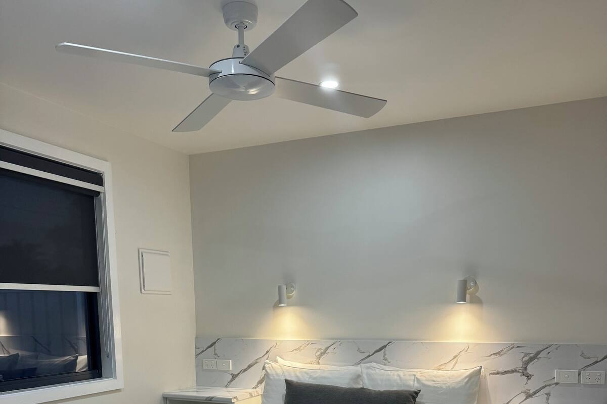 A neatly made bed with a gray and white duvet, a dark gray throw blanket, and two rolled towels. There is a ceiling fan above, two bedside tables with lamps, and a marble-patterned wall.