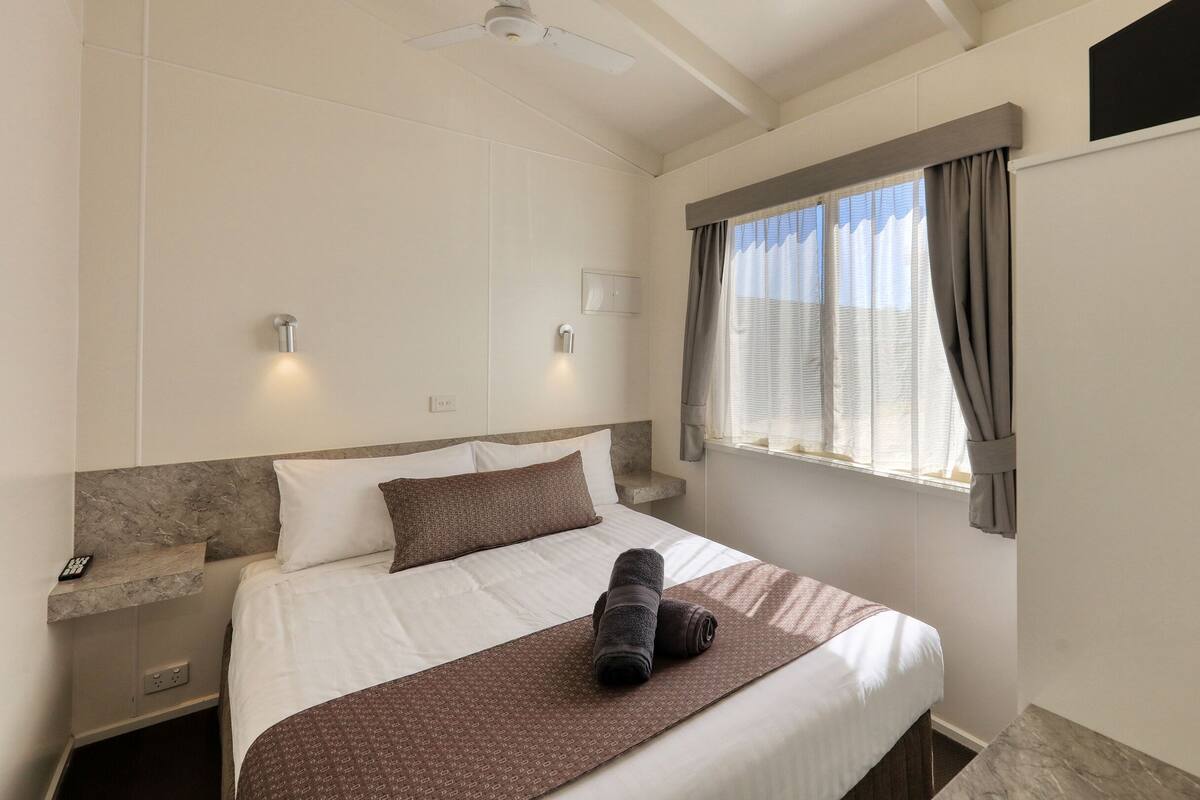 A hotel room with a bed, a nightstand, a wall-mounted light, a window with curtains, and a ceiling fan.