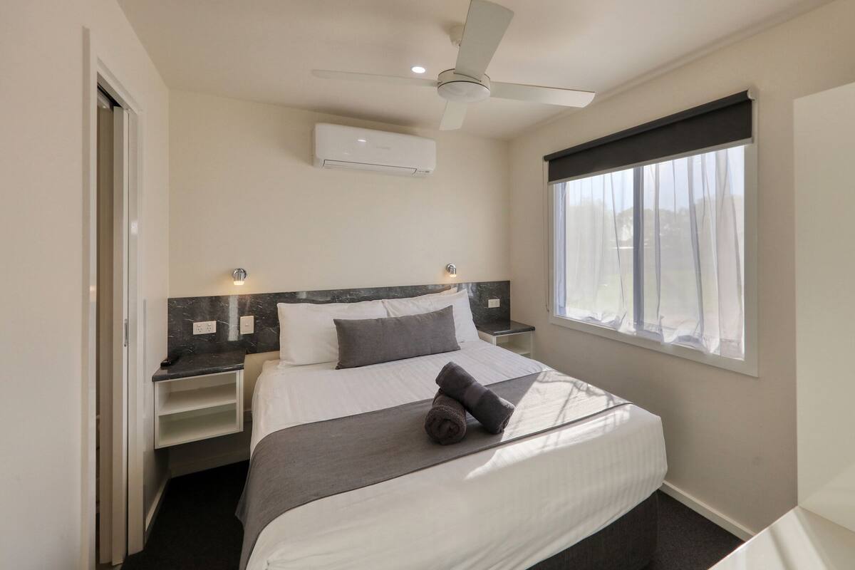 A hotel room with a bed, bedside tables, a ceiling fan, an air conditioning unit, and a window with curtains.