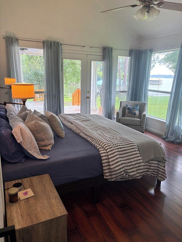 A bedroom with a bed, a chair, a wooden table, and a view of a deck and water outside.