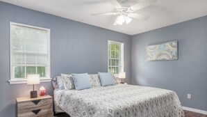 Une chambre à coucher comprenant un lit, deux tables de chevet, une commode, un ventilateur de plafond et deux fenêtres équipées de stores.