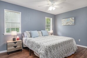 A bedroom with a bed, two bedside tables, a nightstand, a ceiling fan, and two windows with blinds.