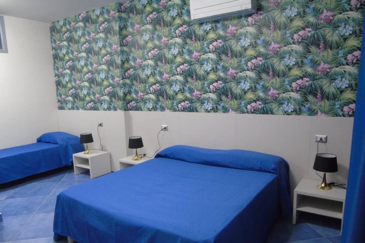 Une chambre avec deux lits, chacun recouvert d’une couverture bleue et agrémenté d’une lampe noire posée sur une table de chevet blanche. Le mur derrière les lits est recouvert d’un papier peint à motifs floraux.