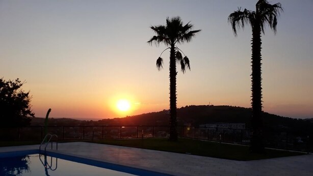 Une piscine au coucher du soleil, avec des palmiers et une vue lointaine sur une colline.