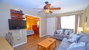 A living room with a sofa, a coffee table, a TV mounted on the wall, a ceiling fan, and a door leading to a bedroom.