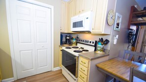 A compact kitchen with a stove, microwave, and a coffee maker. There is a wooden table and a chair, and a small cabinet with drawers.