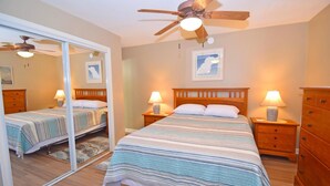 A bedroom with a large bed, two bedside tables with lamps, a wooden dresser, a ceiling fan, and a large mirror.