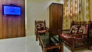 Un salon doté d’un téléviseur à écran plat fixé à un mur en bois, d’un ventilateur de plafond et de deux fauteuils en bois richement décorés, recouverts d’un tissu rouge et or.
