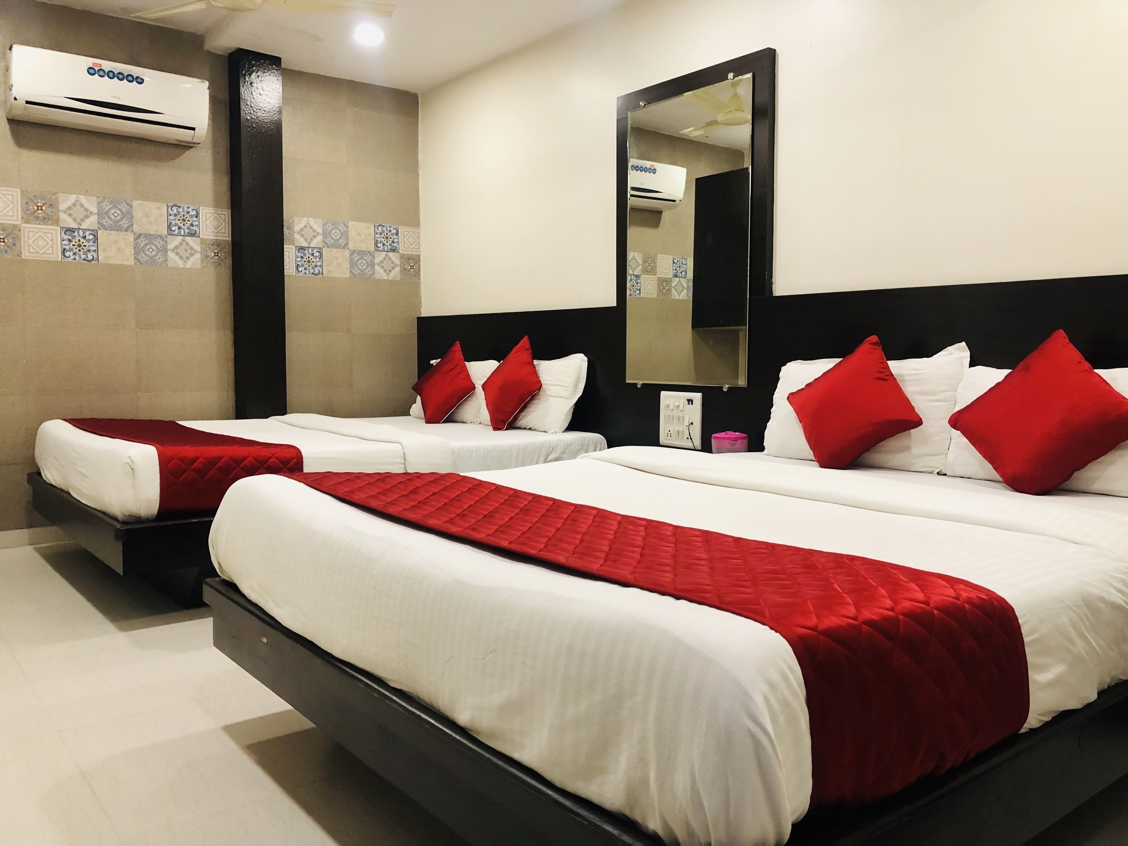 A hotel room with two beds, each with white bedding and red accents, a large mirror, and an air conditioning unit on the wall.