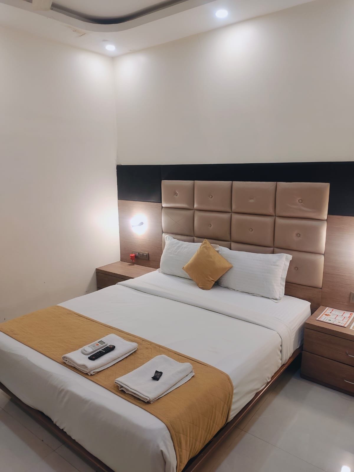 A hotel room with a bed, bedside tables, a nightstand, and a wall-mounted light fixture.