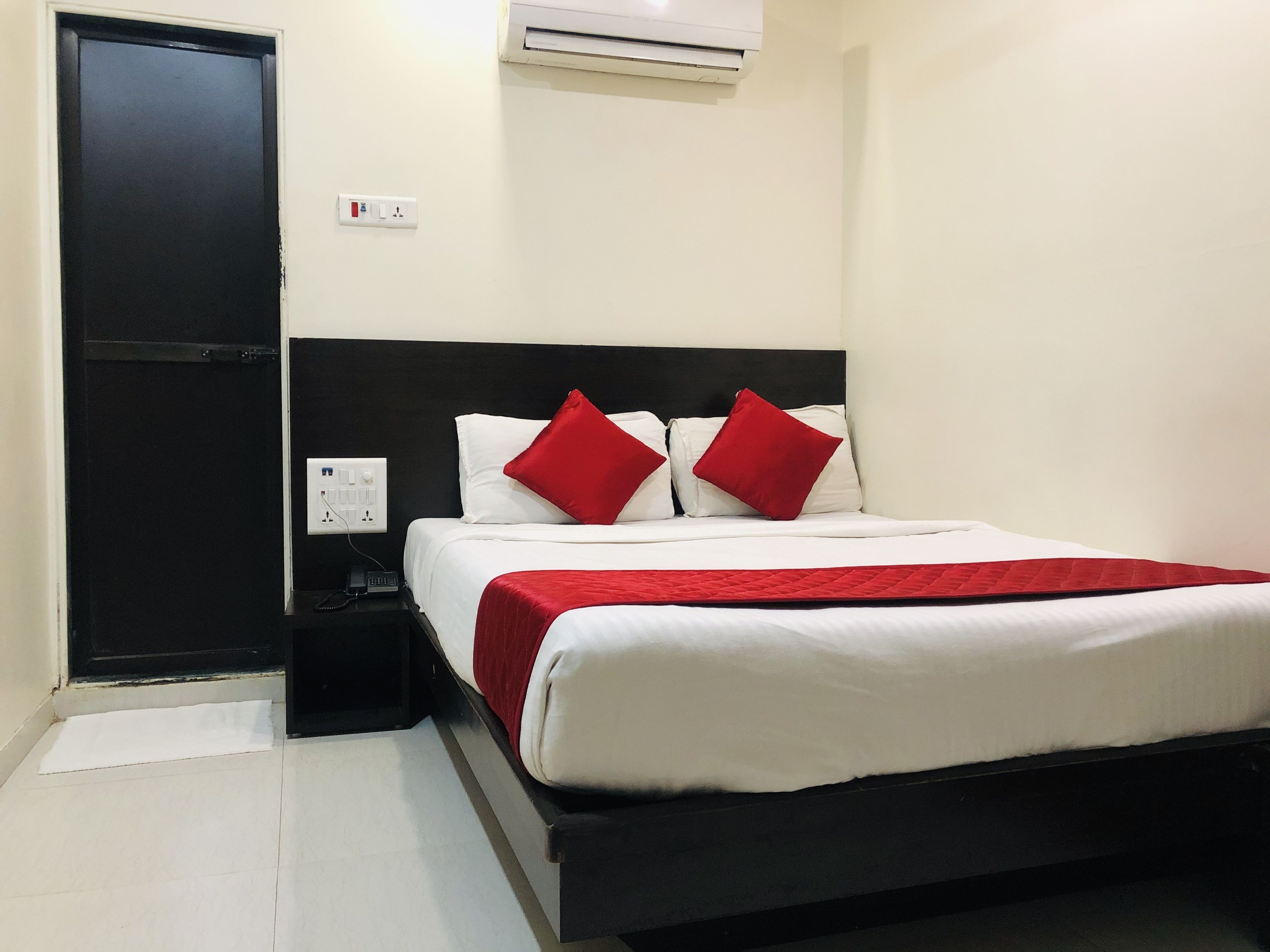 A hotel room with a bed, a nightstand, a power outlet, and an air conditioning unit.