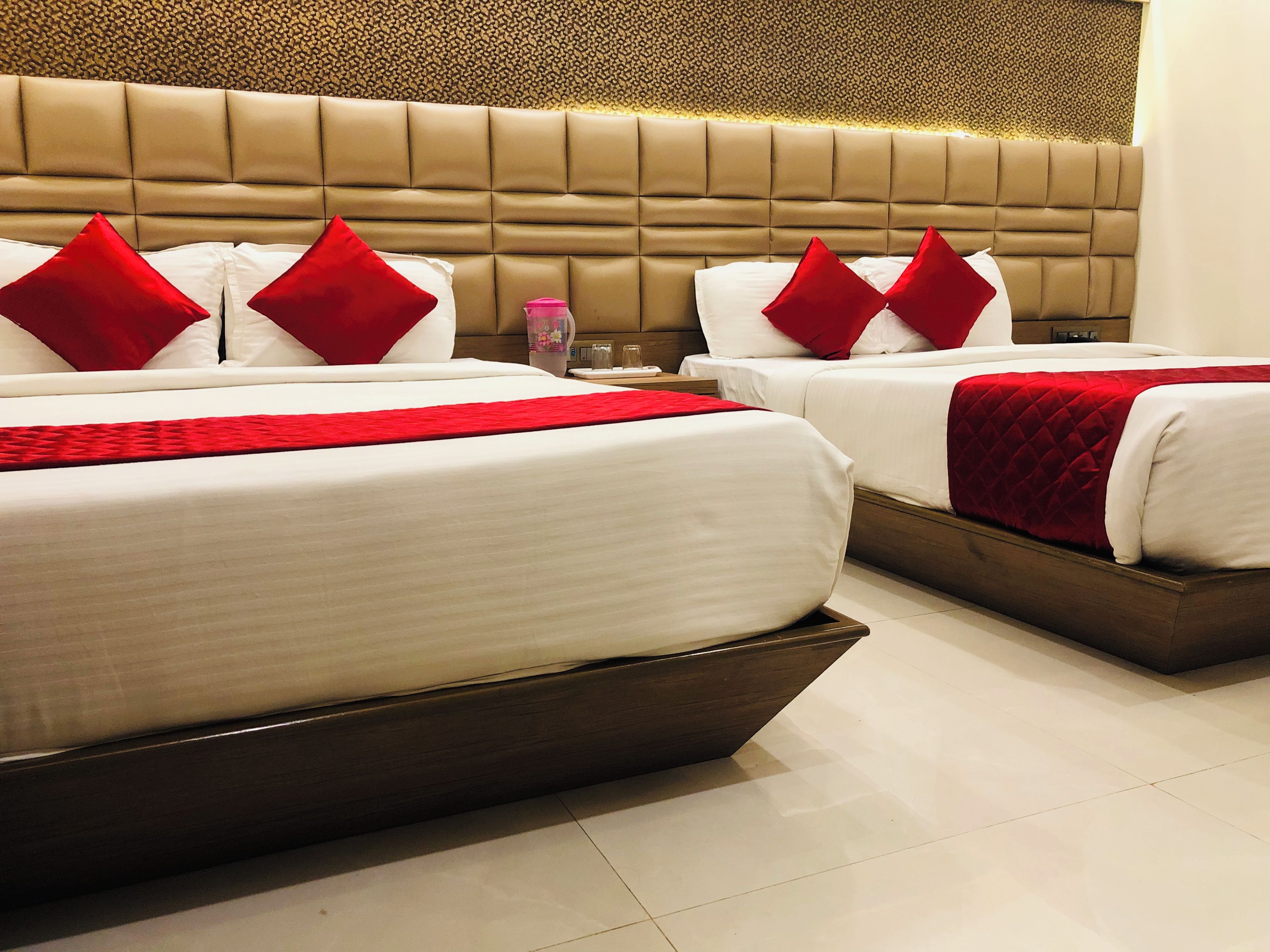 Une chambre d’hôtel avec deux lits, chacun doté de draps blancs et d’oreillers rouges, d’une grande tête de lit rembourrée et d’un sol carrelé.