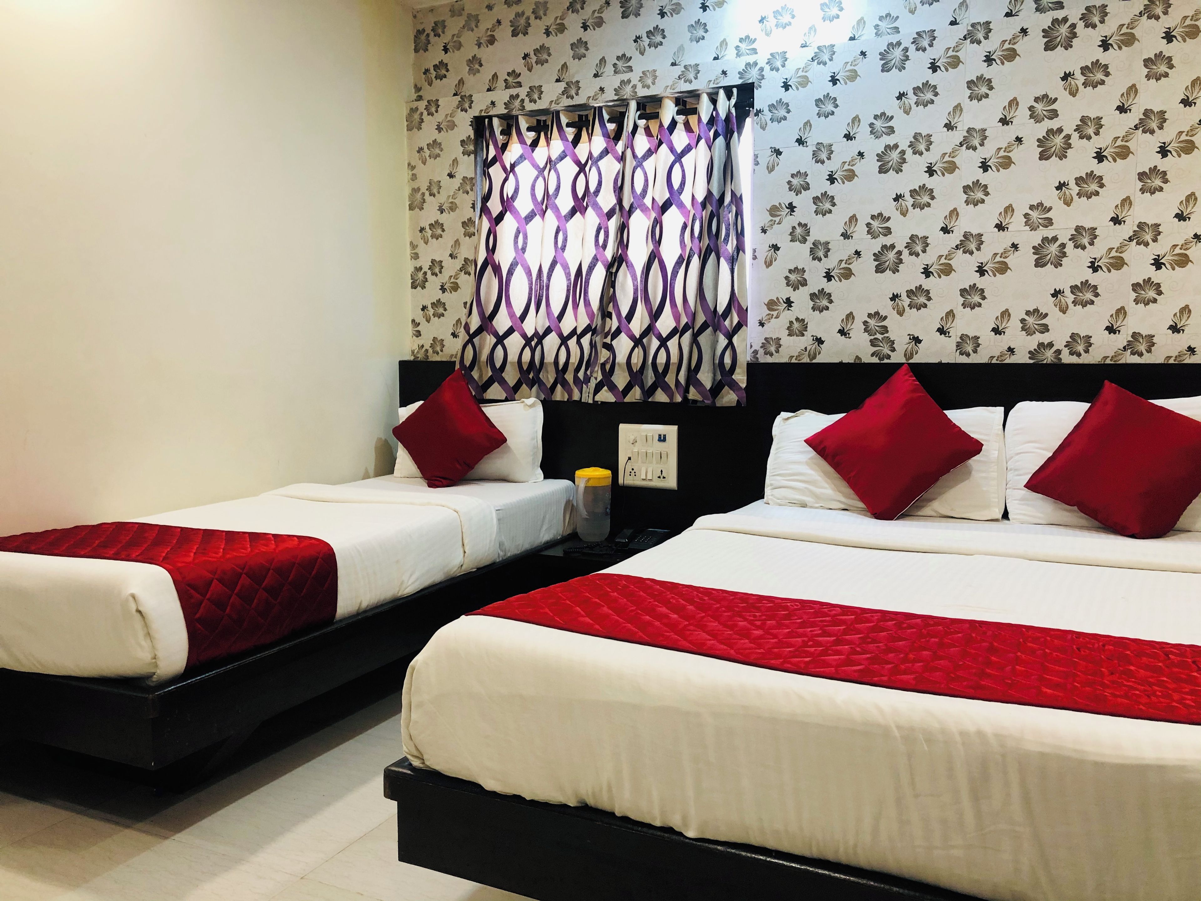 A hotel room with two beds, each with red and white bedding, a black headboard, and a floral-patterned wall.
