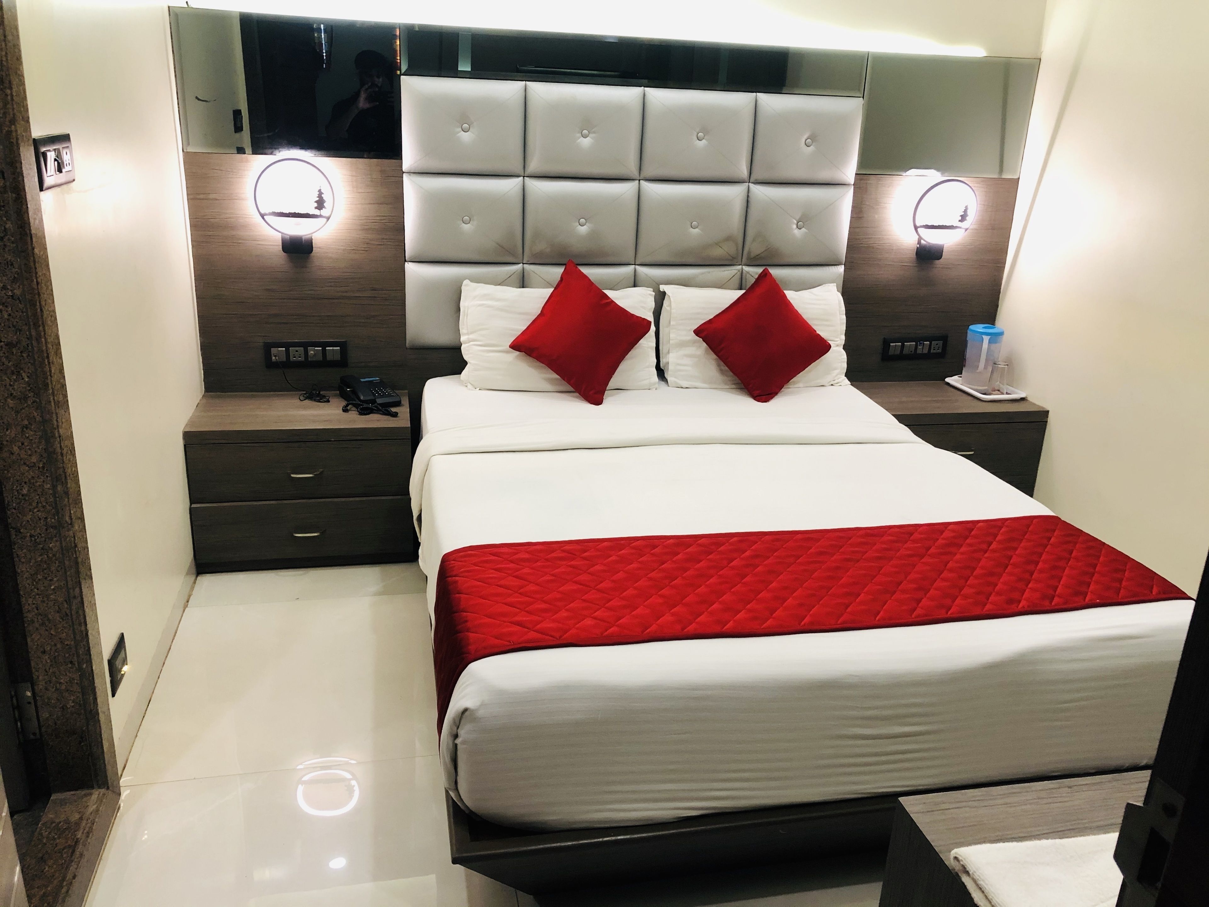 A modern hotel room with a large bed, two bedside tables, a nightstand, and a headboard with built-in lighting.