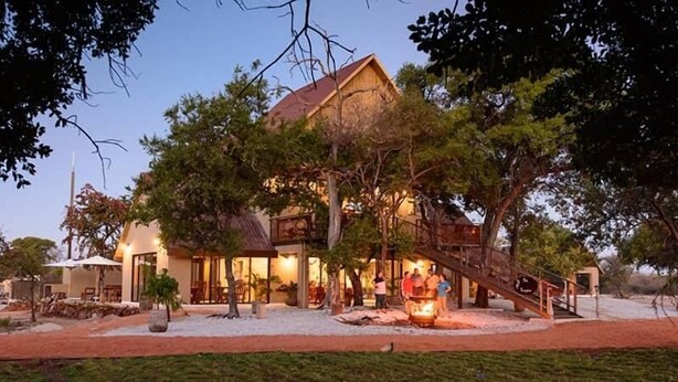 A large, two-story building with a wooden exterior and a balcony. There is a staircase leading to the upper level, and a small fire pit is visible in the foreground. - Callies Game Lodge Safaris (Tsumeb)