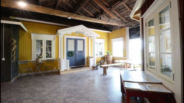 A spacious room with a high ceiling, yellow walls, and a stone floor. There is a wooden bench, a table, and a door with a wreath. - Nygården Bed&Breakfast (Harmanger)