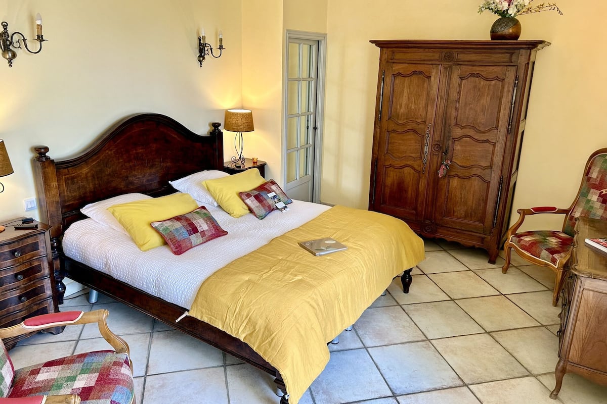 Una cama bien hecha con una colcha amarilla, cabecera de madera y varias almohadas. Se observa un armario de madera con un jarrón de flores encima, una silla con un cojín de colores y una lámpara sobre una mesita de noche.