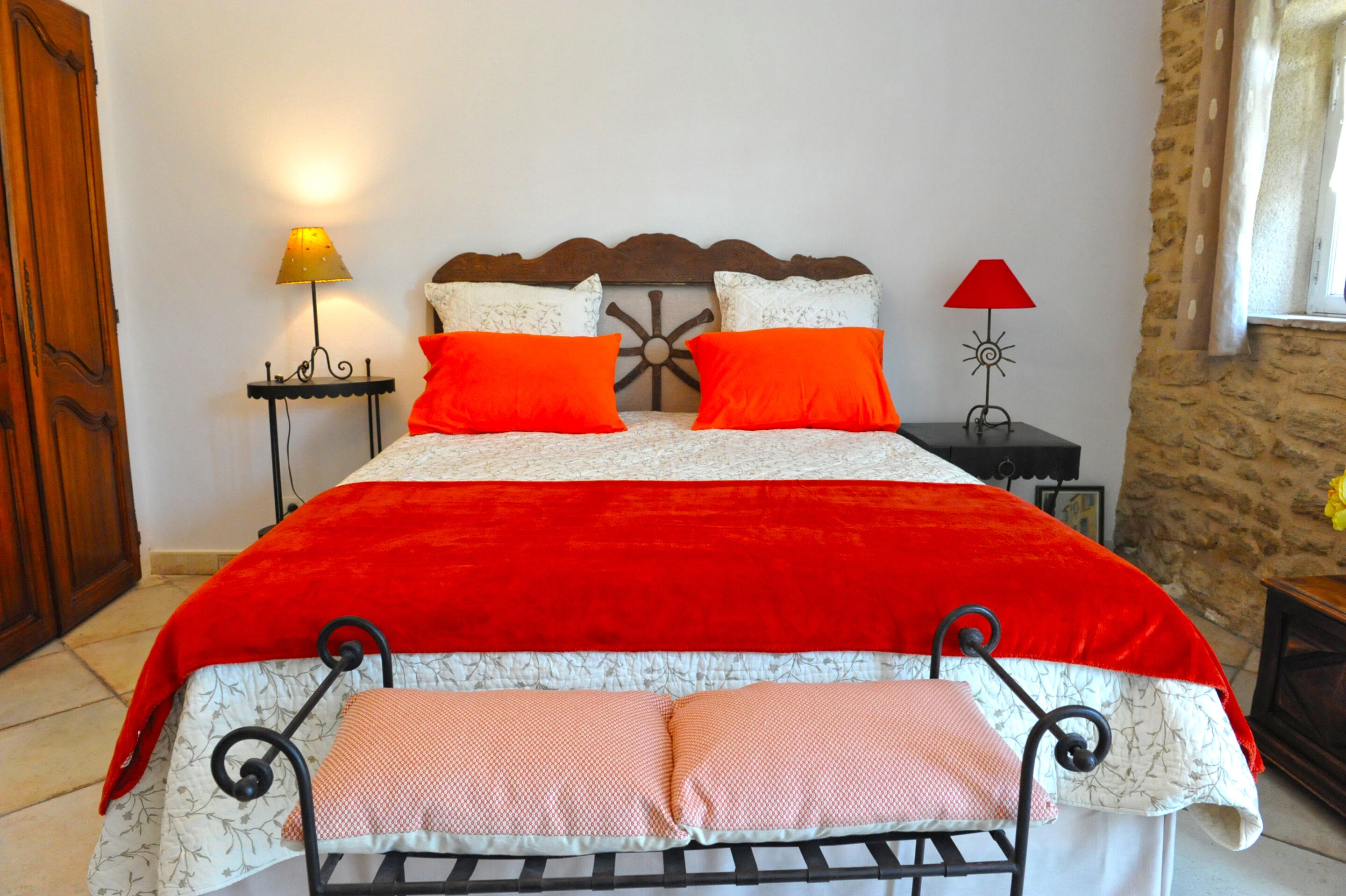 Una cama con una colcha roja y dos almohadas naranjas, un cabecero de madera y dos mesitas de noche con lámparas.