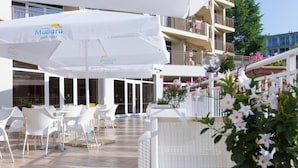 Een terras buiten met witte stoelen en tafels, een grote parasol en een bloeiende plant in een pot.